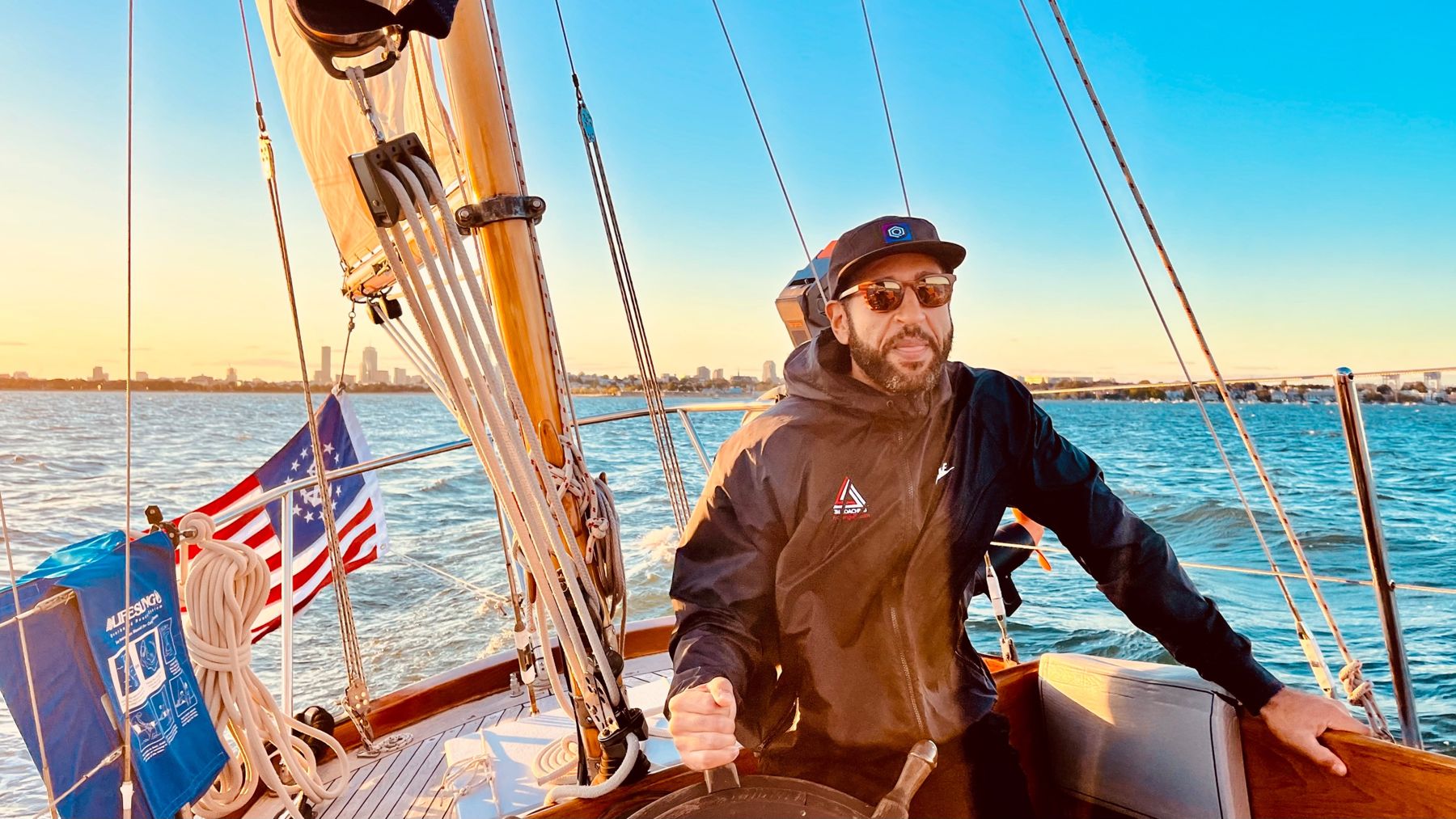 Ian Cain at the helm of a sailboat with the Boston skyline behind him